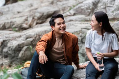 Neşeli bir çift doğa yürüyüşü sırasında kayaların üzerinde oturuyor, gülücüklerini paylaşıyor ve açık hava ortamında iletişim anları kuruyor..