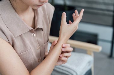 Oturmuş, bileğini rahatsızlıkla tutan bir kadın, sağlık bilincinin ve kişisel bakım uygulamalarının önemini vurguluyor..