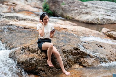 Bir kadın, akmakta olan bir nehrin kenarındaki bir kayanın üzerinde oturmuş müzik dinlerken doğayı kucaklıyor ve dinleniyor..