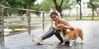Bir kadın yerde oturuyor, Corgi köpeğini severken gülümsüyor, bereketli bir park ortamında evcil hayvan sahibi olmanın sevincini vurguluyor..