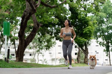 Genç bir kadın, canlı bir parkta Corgi 'siyle koşmaktan zevk alıyor, spor yapmayı ve evcil hayvan arkadaşlığını teşvik ediyor..