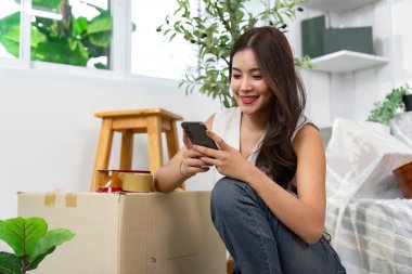 Bir kadın, kutuları taşırken akıllı telefonuna gülümsüyor. Taşınmanın modern deneyimini temsil ediyor..