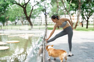 Corgi 'si izlerken bir kadın bacağını uzatıyor. Açık havada sakin bir ortamda spor ve evcil hayvan arkadaşlığının önemini vurguluyor..