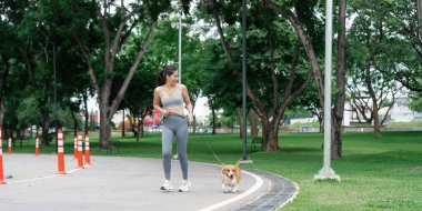 Genç bir kadın, Corgi köpeğini park yolunda gezdirir, sağlığına ve aktif bir yaşam tarzına bağlılığını gösterir..