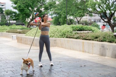 Aktif giyimli neşeli bir kadın, Corgisiyle verimli bir parkta egzersiz yapmaktan zevk alır. Sağlığını ve aktif bir yaşam tarzını destekler..