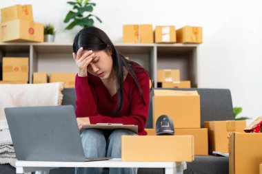 Bir kadın, ev ofisindeki teslimatları yönetirken stresli görünüyor. Uzaktan çalışma ve lojistik zorluklarını yansıtıyor..