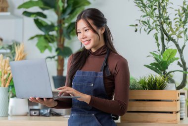 Önlüklü bir kadın ev işleri için dizüstü bilgisayar kullanırken gülümsüyor, teknolojiyi doğayla harmanlıyor..