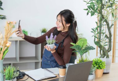 Genç bir kadın bir saksı bitkisiyle selfie çekerken gülümsüyor, modern bir dükkanda bahçe işlerine olan sevgisini gösteriyor..