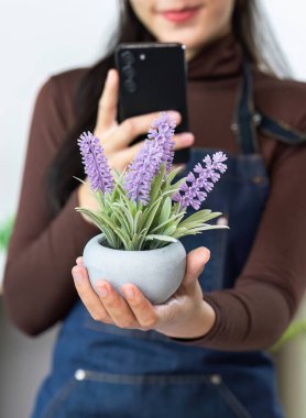 Genç bir kadın, akıllı telefonuyla fotoğraf çekerken saksı çiçeği tutuyor ve bahçe işlerine olan tutkusunu vurguluyor..