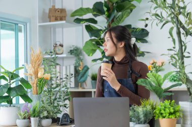Genç bir kadın elinde kahve fincanı tutarken rahat bir dükkanda bitkilere hayranlık duyuyor, sürdürülebilir bir yaşam tarzını somutlaştırıyor..