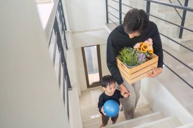 Bir baba ve oğul, merdivenlerden inerken, saksı bitkileri ve bir balo taşırken, aile eğlencesi ve birlikteliği ön plana çıkarırken neşeli bir anı paylaşıyorlar..