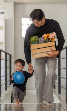 Bir baba ve oğul birlikte yürür, baba çiçekleri taşır ve çocuk mavi bir balon tutar, aile sevgisini ve bağlantısını gösterir..