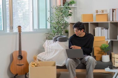 Genç bir adam dizüstü bilgisayarıyla çalışıyor. Yeşillik ve gitarla çevrili şık bir ev ofisinde. Modern bir yaşam tarzı sergiliyor..