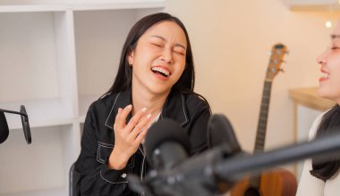 İki kadın podcast yaparken neşeli bir anı paylaşıyor, samimi bir stüdyo ortamında bağlantı ve yaratıcılığı vurguluyor..