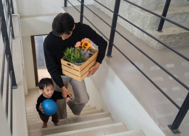 Bir baba oğlunu merdivenlerden indirirken yanında bir sandık çiçek taşır. Aile desteğini ve birlikteliği vurgular..