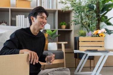 Neşeli genç bir adam modern bir oturma odasında, elinde bir bardak su, rahat ve sağlıklı bir yaşam tarzını somutlaştırıyor..