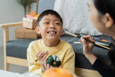 Genç bir çocuk Cadılar Bayramı 'nda annesinin rehberliğinde bir balkabağı çizerken gülümsüyor..