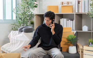 Genç bir adam rahat bir ofiste oturuyor, telefonla konuşuyor ve çalışma alanını kutular ve bitkilerle yönetiyor..