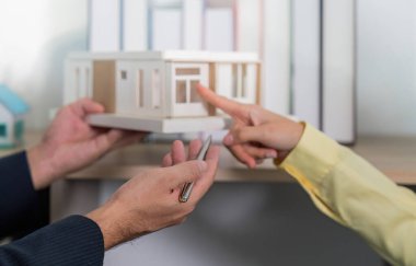 Müşteri, modern ofis ortamında bir emlakçıyla mimari ayrıntıları tartışırken model bir evi işaret eder..