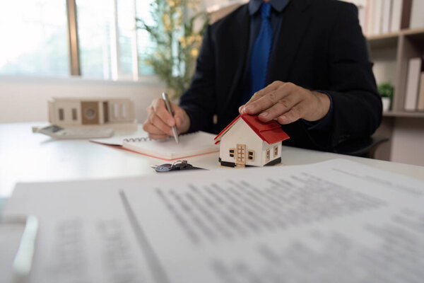 A real estate agent takes notes while examining a small house model, preparing for a client meeting in a modern office.