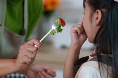 Bir ebeveyn çocuğuna taze malzemelerle dolu neşeli bir mutfak ortamında sağlıklı beslenme alışkanlıklarını teşvik etmek için bir parça salata sunar..