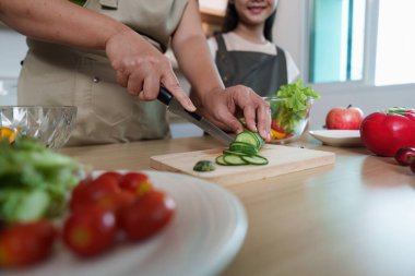 Bir anne, kızı izlerken salatalıkları dilimlerken sağlıklı beslenme alışkanlıklarının önemini vurgular..