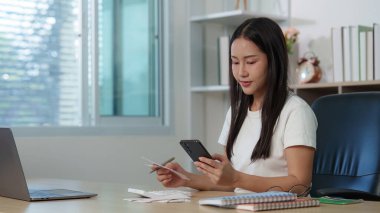 Akıllı telefonunu kullanarak finansal belgeleri inceleyen genç bir kadın, etkili bütçe uygulamalarını vurguluyor.