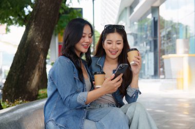 İki arkadaş neşeli bir anı paylaşıyor. Akıllı bir telefona bakıp, kahvelerini yudumlarken, açık havada canlı bir mekandaki bağlantılarını vurguluyorlar..