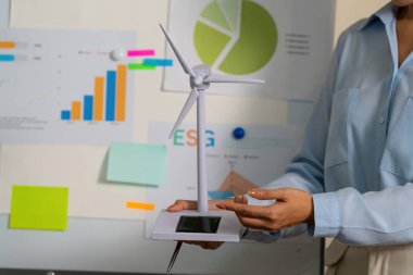 Bir iş kadını ESG sunumunda yenilenebilir enerji çözümlerini vurgulayan bir rüzgar türbini modeli tutuyor.