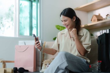 Genç bir kadın, yanında kedisi olan akıllı telefonuyla çevrimiçi siparişlerini kontrol ederken gülümsüyor. E-ticaretin neşesini temsil ediyor.