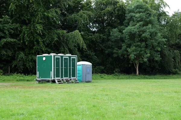 Four portable toilets have been setup at the edge of a playing field, in front of trees. One of the toilets  has been designated as an accesible one.