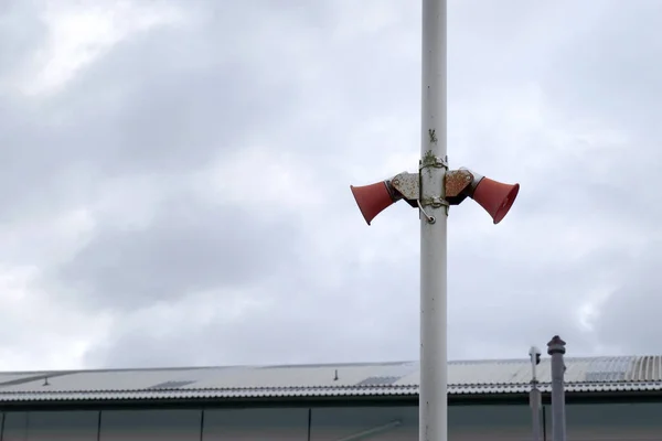 İki kırmızı, biraz paslı megafon claxon beyaz metal bir direğe tutturulmuş. Arka planda bir indsutrial birim görülebilir.