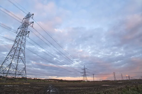 Wide angle of electrical power lines at dusk with a blue and pink sky. The lines lead away across a muddy field, to a vanishing point in the distance.