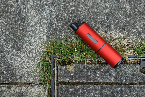 Tek kullanımlık, tek kullanımlık elektronik bir sigara kabı, yanında yabani otlar büyüyen gri betonun üzerine bırakılmış..