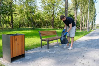 Bir erkek gönüllü halka açık bir parkta siyah bir çantada plastik atık topluyor. Çevrenin temizliği. Çevresel sorunlar