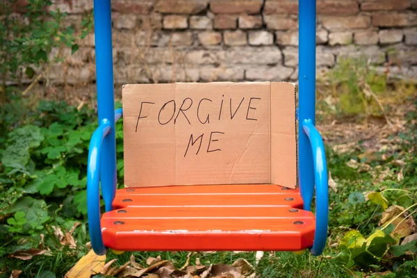 Text with the inscriptions Forgive me, handwritten on cardboard on a children\'s swing. Please forgive me. Forgiveness concept