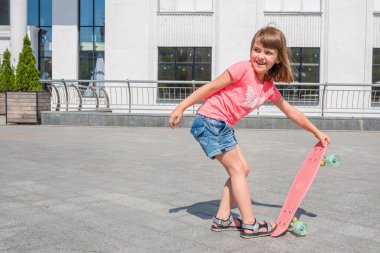 Güneşli bir yaz gününde, nehir kıyısındaki parkta kaykaylı mutlu küçük bir kızın açık hava portresi..