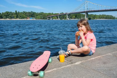 Küçük bir kız şehir nehir arka planında hamburger yiyor. Yaşam tarzı. Aç bebek..