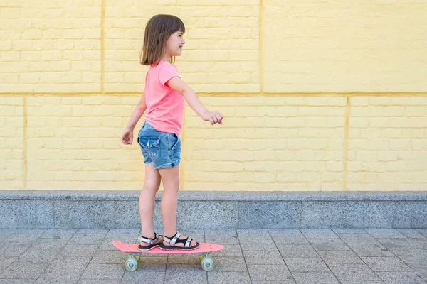 ピンクのTシャツの7 8歳の少女は 黄色い壁の背景でスケートボードをしています ライフスタイル — ストック写真