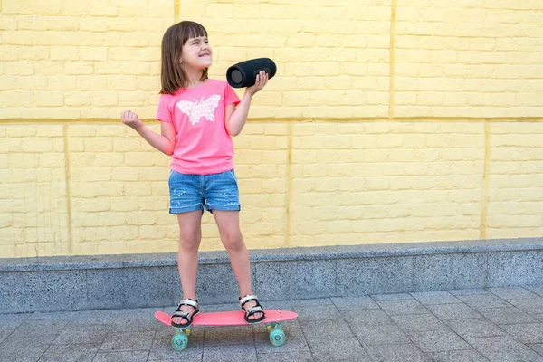 音楽ブルートゥーススピーカーを持つ小さなスタイリッシュな少女は 黄色いレンガの壁の背景でスケートボードをしています ライフスタイル — ストック写真