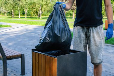 Gönüllü bir adam, şehir parkındaki bir çöp kutusuna siyah bir çanta atıyor. Sokak temizliği, çevre sorunları. Çevre koruma kavramı