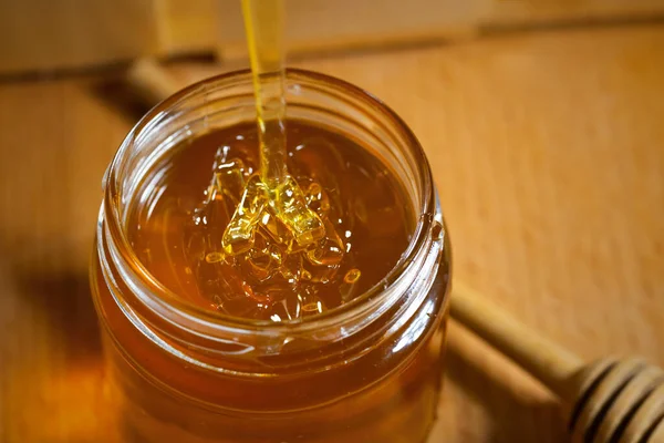 Pouring aromatic honey into jar, closeup. A glass jar of honey and a wooden honey dipper on a wooden background.