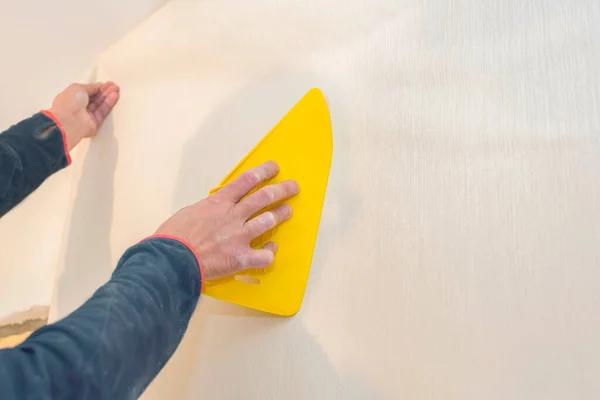 İşçi, duvar kağıdını plastik sarı bir spatulayla yumuşatıyor. Onarım kavramı.