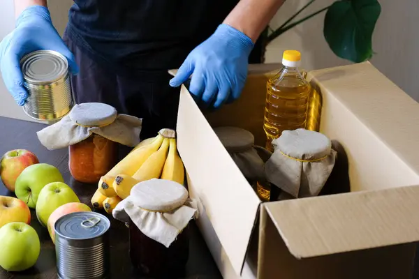 A volunteer in protective gloves lays foods into a box. Delivery during quarantine. Assistance concept during quarantine, charity.