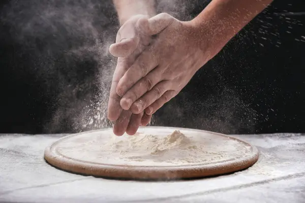 A male chef claps his hands filled with white flour on a black background. Homemade baking process, baking concept, close-up.