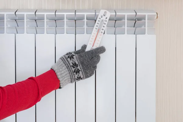 A mans hand in a warm glove holds a thermometer near the radiator. Temperature check, cold winter season.