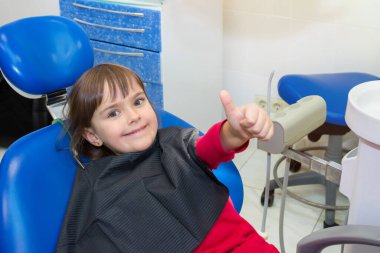 Mutlu bir çocuk dişçi koltuğunda başparmağı havada oturuyor. İlaç, dişçilik ve sağlık hizmetleri.