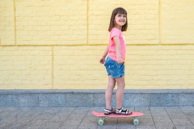 Zarif küçük bir kız sarı duvar arkasında kaykay yapıyor. Spor yaşam tarzı..
