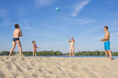 Çocuklar ve aileleri güneşli bir yaz gününde nehir kıyısında voleybol oynarlar..