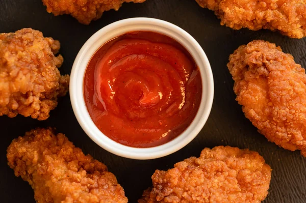 Fried breaded chicken strips with red sauce on a black table. View from above.